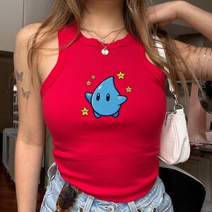 Pu&ograve; includere: Canotta rossa con un personaggio a goccia d'acqua blu dei cartoni animati e accenti di stelle gialle. La parte superiore &egrave; in stile a maglia a coste.