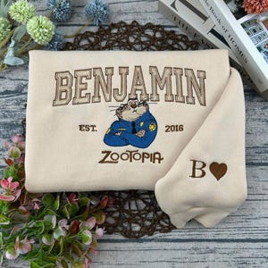 Puede incluir: Sudadera color crema con bordado "BENJAMIN" sobre un personaje de dibujos animados con uniforme de polic&iacute;a. Debajo, "ZOOTOPIA" y "EST. 2016". La manga tiene una "B" y un coraz&oacute;n.