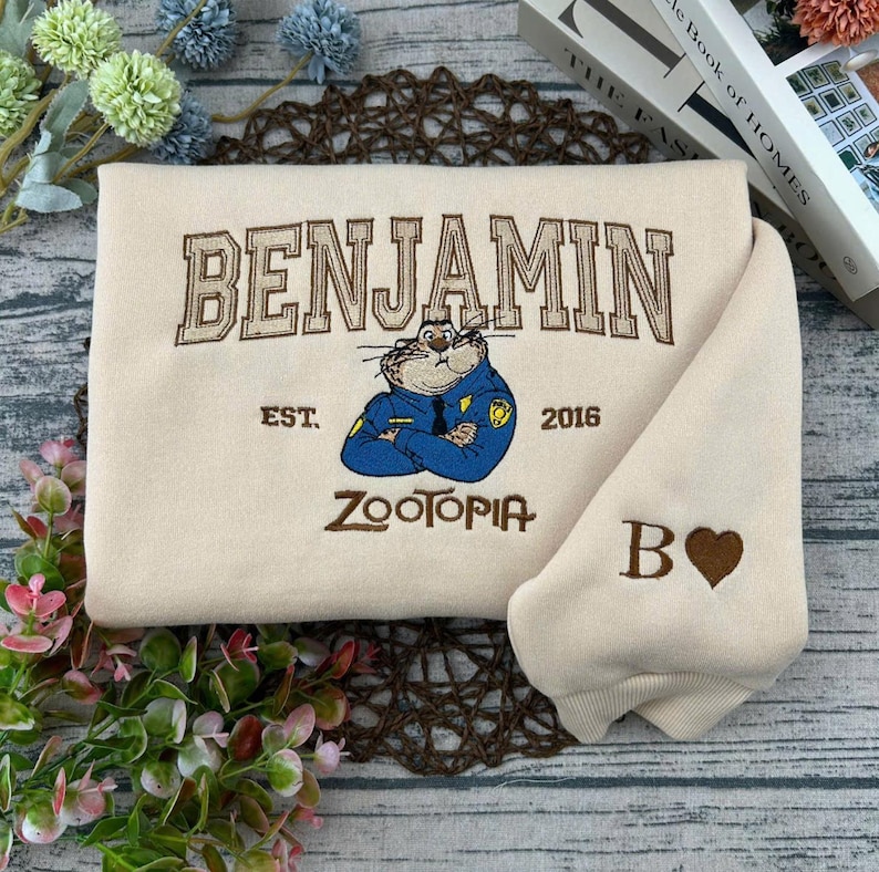 Puede incluir: Sudadera color crema con bordado "BENJAMIN" sobre un personaje de dibujos animados con uniforme de polic&iacute;a. Debajo, "ZOOTOPIA" y "EST. 2016". La manga tiene una "B" y un coraz&oacute;n.
