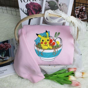 May include: A light pink sweatshirt featuring an embroidered design of Pikachu in a bowl of ramen. The bowl is blue and white, and the ramen includes noodles, an egg, and other ingredients. The sweatshirt is folded in a basket with magazines and flowers.