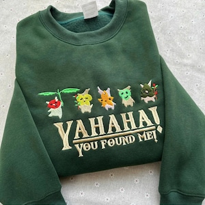 Könnte beinhalten: Dunkelgrünes Sweatshirt mit gestickten Cartoon-Figuren und dem Text "YAHAHAI YOU FOUND ME!". Das Design zeigt farbenfrohe, skurrile Figuren mit pflanzenartigen Merkmalen. Das Sweatshirt hat einen Rundhalsausschnitt und lange Ärmel.