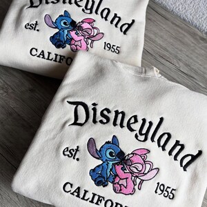 May include: Two cream-coloured sweatshirts with the embroidered text "Disneyland" and "CALIFORNIA 1955". Each sweatshirt features a blue and pink cartoon character kissing. The sweatshirts are made of a soft material.