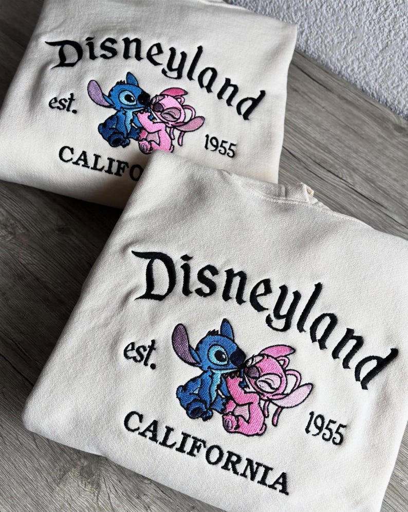 May include: Two cream-coloured sweatshirts with the embroidered text "Disneyland" and "CALIFORNIA 1955". Each sweatshirt features a blue and pink cartoon character kissing. The sweatshirts are made of a soft material.