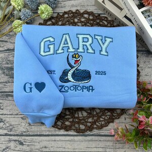Puede incluir: Sudadera azul claro con el nombre "GARY" en letras azul claro. Tiene un dise&ntilde;o de serpiente bordada y la palabra "ZOOTOPIA". La manga muestra la letra "G" y un coraz&oacute;n azul.