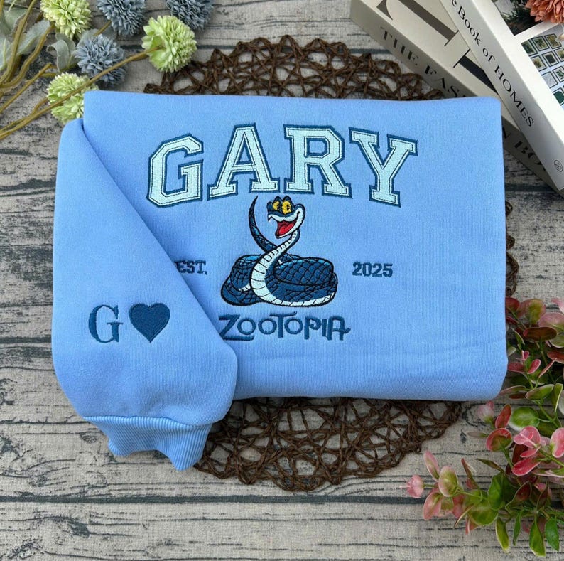 Puede incluir: Sudadera azul claro con el nombre "GARY" en letras azul claro. Tiene un dise&ntilde;o de serpiente bordada y la palabra "ZOOTOPIA". La manga muestra la letra "G" y un coraz&oacute;n azul.