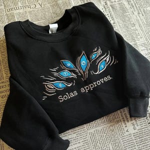 May include: Black sweatshirt with embroidered design featuring multiple blue eyes and the text "Solas approves." The design is in shades of blue and white. The sweatshirt is made of a soft material.