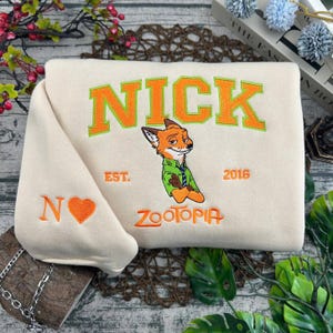 Puede incluir: Sudadera color crema con el nombre "NICK" en letras naranja con borde verde. Un personaje de zorro bordado de Zootopia est&aacute; debajo del nombre, con el texto "EST. 2016" y "ZOOTOPIA". La manga tiene una "N" bordada y un coraz&oacute;n.
