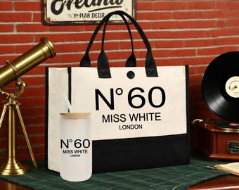 Op de afbeelding: Een crèmekleurige canvas draagtas met zwarte handvatten en een zwart paneel, bedrukt met "N°60 MISS WHITE LONDON". Een glazen beker met bamboe deksel en rietje, met dezelfde tekst, staat voor de tas.