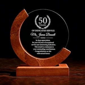 May include: A clear glass award with a wooden base and a curved wooden accent. The award features the text "50 YEARS OF DEDICATED SERVICE" and a personalized message. The award is designed to honor 50 years of service.