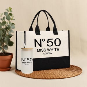 Può includere: Una borsa tote bianca con manici neri e base nera. La borsa presenta la scritta "N° 50 MISS WHITE LONDON" in nero. Un bicchiere di vetro smerigliato con coperchio e cannuccia in bambù è davanti, con lo stesso testo.
