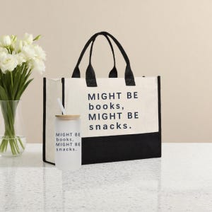 Może przedstawiać: Płócienna torba z czarnymi uchwytami i czarnym dolnym panelem. Torba ma napis "MIGHT BE books, MIGHT BE snacks." Przed torbą znajduje się szklany kubek z matowego szkła ze słomką i tym samym tekstem.