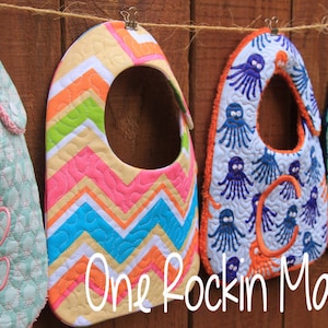 May include: Four colourful baby bibs hanging on a wooden fence. The bibs are made of fabric and have different patterns, including a chevron pattern, an octopus pattern, and an anchor pattern. The bibs are all personalised with embroidered letters.