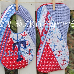 May include: Two red, white, and blue baby bibs with an anchor and wave pattern. One bib has the letter "T" and the name "thomas" embroidered on it. The bibs are hanging on a white picket fence.