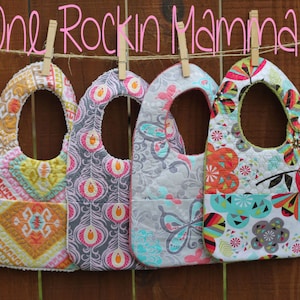 Può includere: Quattro bavaglini per neonati colorati con diversi motivi appesi a uno stendino. I bavaglini sono realizzati in tessuto trapuntato e hanno una tasca sul fondo. Il testo "One Rockin Mamma" è scritto sopra i bavaglini.