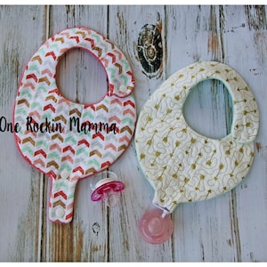 May include: Two baby bibs with a geometric arrow pattern. One bib is pink, white, and gold with a pacifier holder. The other bib is light blue and gold with a pacifier holder. The text "One Rockin Mamma" is visible in the image.