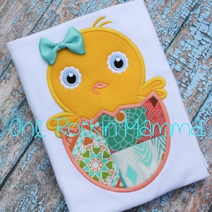 May include: A yellow chick with a blue bow peeks out of a colourful patchwork egg. The chick has white eyes and a pink beak. The egg is surrounded by a pink outline.