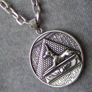 Puede incluir: Collar con colgante plateado con un diseño de temática egipcia. El colgante redondo muestra una pirámide, jeroglíficos y una figura de chacal reclinada. El collar tiene una cadena con eslabones ovalados.