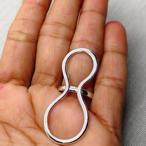 Puede incluir: Un anillo plateado con un diseño único en forma de ocho. El anillo está hecho de metal pulido y presenta una forma de bucle continuo. El anillo se muestra en una mano con un fondo neutro.