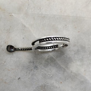 May include: A silver ring with a rope-like design around the band. A small, decorative spoon-like element is attached to the ring. The ring is set against a light-colored, textured background.