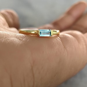 Peut inclure: Une bague en or ornée d'une unique pierre précieuse bleu clair de taille baguette rectangulaire, montée horizontalement dans un serti clos. L'anneau est lisse et poli, présentant un design simple qui met en valeur la clarté de la pierre.