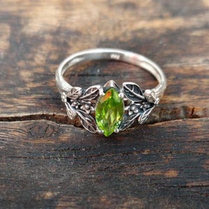 May include: A silver ring with a light green, marquise-cut gemstone. The ring has a floral design with leaves and small flowers on the band. The gemstone is the focal point, set in a detailed, nature-inspired setting.
