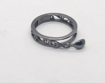 May include: A dark gray ring with an openwork design and a small, teardrop-shaped charm. The ring features a decorative, swirling pattern around the band. The charm hangs from the ring's side.