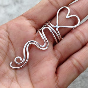 Op de afbeelding: Een zilverkleurige ring met een hartvormig ontwerp en een golvend, abstract patroon. De ring heeft een dubbelbandig, golvend ontwerp dat om de vinger gewikkeld is. De hartvorm bevindt zich bovenaan de ring.