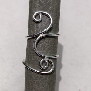 May include: A silver-colored ring with a unique design. The ring features a swirling, openwork pattern with two spiral accents. The ring is shown on a gray surface, highlighting its intricate details and modern aesthetic.
