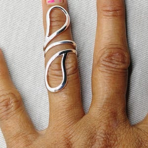 Puede incluir: Un anillo plateado con un diseño abierto, con dos elementos en forma de lágrima conectados por una banda. El anillo se lleva en un dedo, mostrando su estilo único y moderno. La superficie lisa y reflectante del anillo captura la luz.