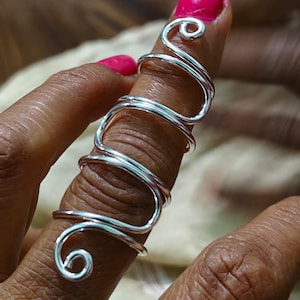 Könnte beinhalten: Ein silberfarbener Drahtring mit einem einzigartigen, fließenden Design. Der Ring umschließt den Finger mit einer Reihe von Kurven und Wirbeln und schafft so ein auffälliges Accessoire. Der Ring wird auf einem Finger mit leuchtend pinkfarbenem Nagellack gezeigt.