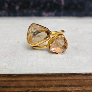 Könnte beinhalten: Ein goldfarbener Bypass-Ring mit zwei rohen, durchscheinenden, hell bernsteinfarbenen Edelsteinen. Die Steine sind unregelmäßig geformt, haben facettierte Kanten und sind in ein glattes, poliertes, goldfarbenes Metallband eingefasst.