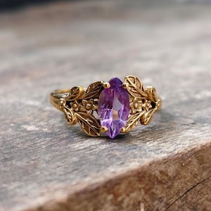 Op de afbeelding: Een goudkleurige ring met een paarse marquise-geslepen edelsteen. De ring is versierd met een blad- en bloemmotief rond de edelsteen, wat een door de natuur geïnspireerde esthetiek creëert. De ring ligt op een houten oppervlak.