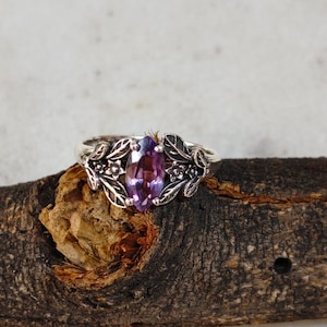 Op de afbeelding: Een zilveren ring met een marquise-geslepen paarse edelsteen, omgeven door een gedetailleerd bloemen- en bladerontwerp. De ring is geplaatst op een rustiek houten oppervlak, dat het ingewikkelde metaalwerk en de levendige kleur van de steen benadrukt.
