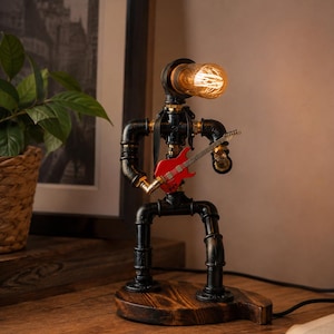 May include: A table lamp constructed from black industrial metal pipes, shaped like a figure playing a red electric guitar. The head of the figure is a glowing Edison-style light bulb. The entire structure is mounted on a circular wooden base.