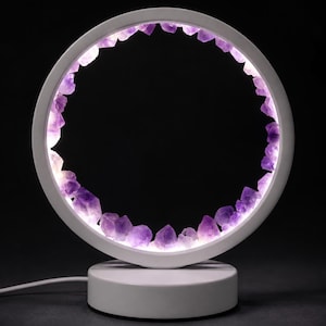 May include: A circular white table lamp featuring a ring frame lined with purple amethyst crystal clusters. The interior of the ring is illuminated by integrated LED lights, casting a glow through the translucent purple stones. The lamp sits on a round white base.