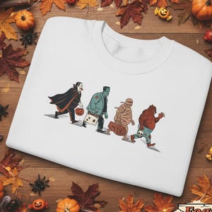 Könnte beinhalten: Weißes Sweatshirt mit Rundhalsausschnitt und Halloween-Motiv. Die Grafik zeigt Dracula, Frankensteins Monster, eine Mumie und einen Werwolf, jeder mit einem Trick-or-Treat-Beutel. Der Hintergrund ist Holz, mit Kürbissen und Herbstblättern.