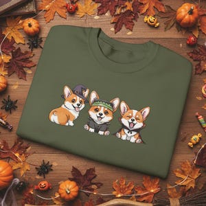 Może przedstawiać: Oliwkowo-zielona bluza z trzema kreskówkowymi corgi w kostiumach na Halloween. Jeden nosi kapelusz czarownicy, drugi kostium Frankensteina, a trzeci pelerynę wampira. Obraz otoczony jest jesiennymi liśćmi i dyniami.