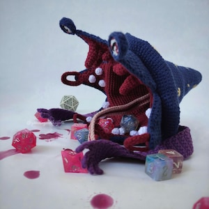 May include: A handmade crochet monster hat in navy blue and burgundy, with white detailing. The hat is surrounded by colourful polyhedral dice, with some scattered on the surface. The hat has a unique, tentacled design, perfect for tabletop gaming.