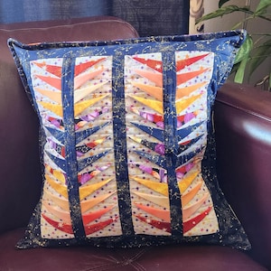 May include: A square decorative pillow with a quilted design. The pillow features a dark blue border with gold star and constellation accents. The center has vertical panels of colorful, layered triangles in shades of orange, yellow, and purple.