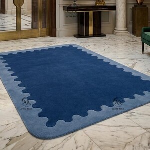Op de afbeelding: Een rechthoekig vloerkleed met een diepblauw midden en een lichtblauwe geschulpte rand. Het kleed ligt op een witte marmeren vloer in een grote hal met een kristallen kroonluchter, een sidetable en groene bankjes.