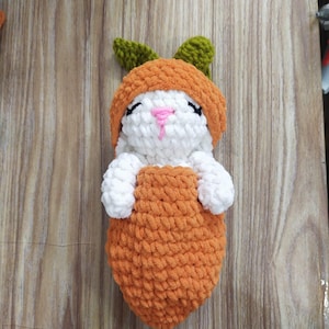 May include: A handmade crochet bunny doll in a carrot costume. The bunny is white with a pink nose and black eyes. The carrot costume is orange with green leaves on top. The doll is lying on a wooden surface.