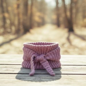 May include: A mauve-colored, crocheted neck warmer with a bow detail. The handmade cowl is made of a thick yarn and sits on a wooden surface. The background is blurred, showing a path through a forest.