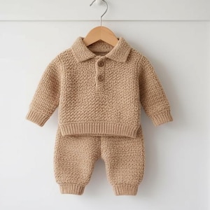 May include: A tan knitted baby outfit, comprising a long-sleeved polo shirt and matching trousers, hangs on a wooden hanger. The shirt features a collared neckline and two buttons. The outfit is made of a textured knit fabric.