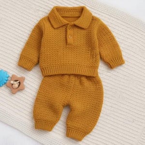 Pode incluir: Conjunto de bebé em malha amarelo mostarda, composto por uma camisola de manga comprida com gola e dois botões e calças a condizer. O conjunto é feito de tecido de malha texturizado. O conjunto é apresentado numa manta macia de cor neutra.
