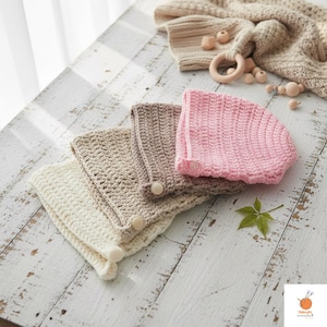 May include: A collection of handmade crochet baby bonnets in cream, tan, and pink, with a matching blanket and wooden teething ring. The bonnets have a button closure and are arranged on a distressed white wooden surface.