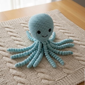 Op de afbeelding: Een lichtblauwe gehaakte octopus met een lachend gezicht en acht tentakels, rustend op een crèmekleurige mat met kabelpatroon. De octopus is handgemaakt en ziet er zacht uit. Het algemene thema is een handgemaakt speelgoed.