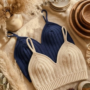 May include: Two handmade crochet tops. One is a light beige colour, and the other is navy blue. Both tops have a V-neck design and thin straps. The tops are displayed on a neutral-coloured surface, with decorative elements in the background.