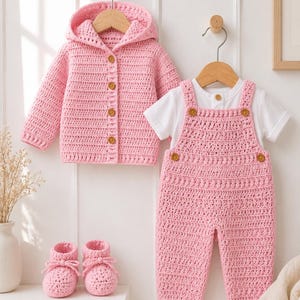 Puede incluir: Un cárdigan con capucha de ganchillo rosa con botones marrones, una camiseta blanca de manga corta, un peto de ganchillo rosa con botones marrones y un par de patucos de ganchillo rosa a juego con cordones, presentados como un conjunto.