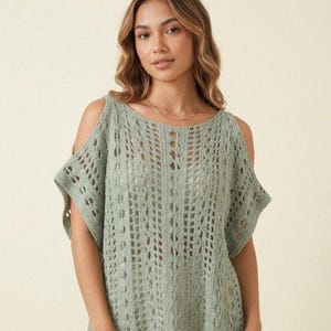Puede incluir: Una blusa de ganchillo color verde salvia con un patrón de tejido abierto, columnas verticales de textura y aberturas en los hombros. La prenda tiene un cuello redondo y mangas cortas, mostrando una construcción tejida detallada y aireada.