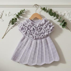 May include: A light purple, handmade crochet dress with a ruffled collar, hanging on a wooden hanger. The dress has a flared skirt and a textured stitch pattern. The background features white wall decor and greenery.